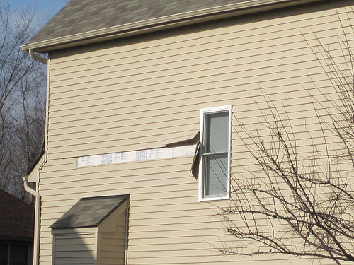 Side of tan home with missing and damaged vinyl siding from a storm.