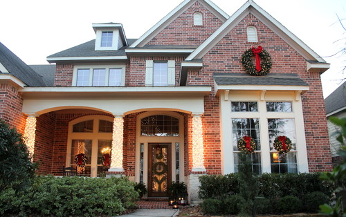 Two story brick home with christmas wreaths on windows and lights covering the front porch columns.