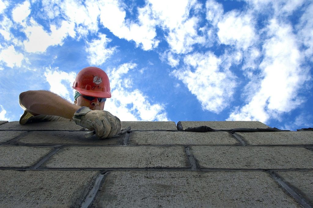 Man with red helmet reaching over the peak of a roof working on shingles.