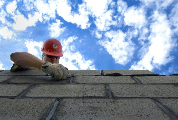 Man with red helmet reaching over the peak of a roof working on shingles.