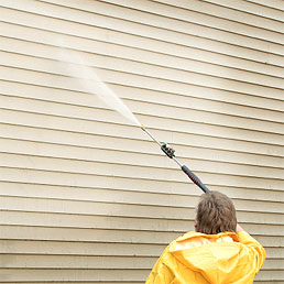 Man in yellow rain coat power washing tan siding on a home.
