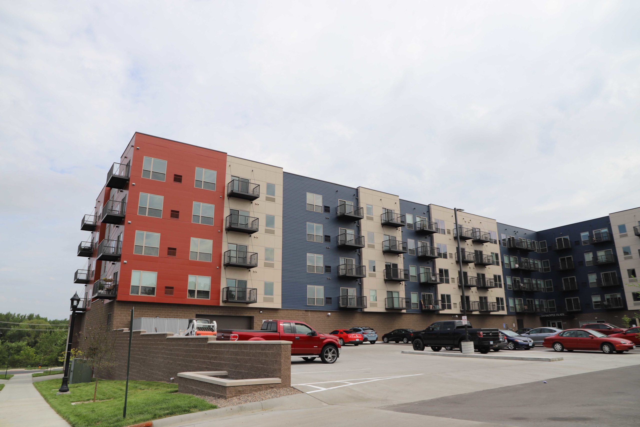 End of a red, cream, and blue, five-story apartment building with a parking lot in Shakopee, MN.