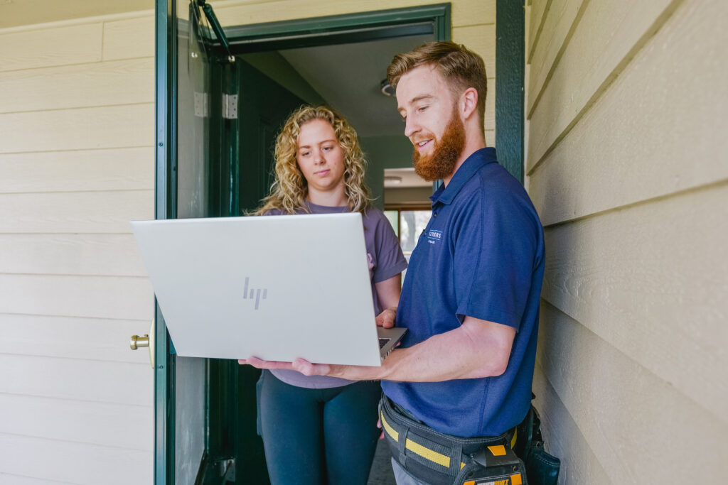 A technician, wearing a blue shirt and tool belt, shows a woman something on a laptop at her doorway. The woman, dressed casually, is attentively looking at the screen. They're likely reviewing a fall home maintenance checklist for homeowners to ensure everything is in order.