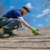 A person wearing a headlamp and gloves inspects the shingles of a roof on a sunny day. They have a tool belt strapped to their waist and are crouching down to closely examine the surface, following essential storm damage inspection steps.