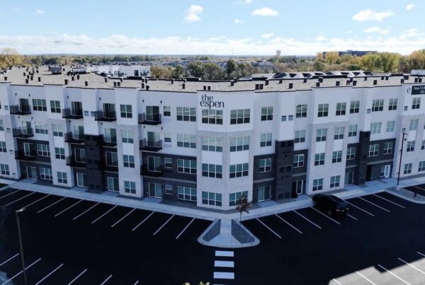 The Espen apartments, a modern multi-story building with balconies, showcases its sleek design against a clear blue sky. On this bright day, the mostly empty parking lot highlights the serene atmosphere surrounding these stylish abodes.