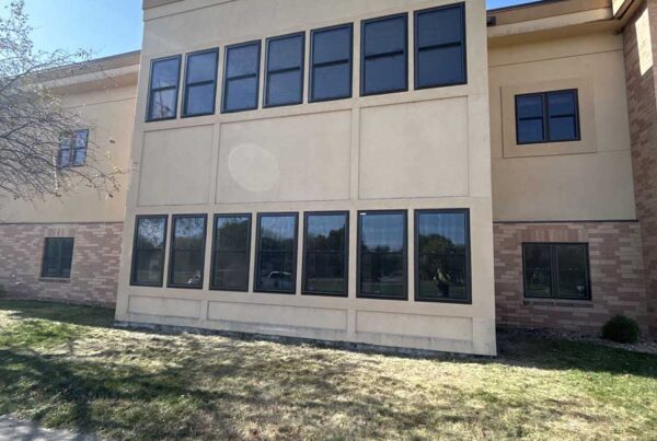 A two-story beige building with tall black-framed windows, brown brick accents at the base, and a clear blue sky in the background—showcasing the results of Oak Hills Living Center Window Replacement.